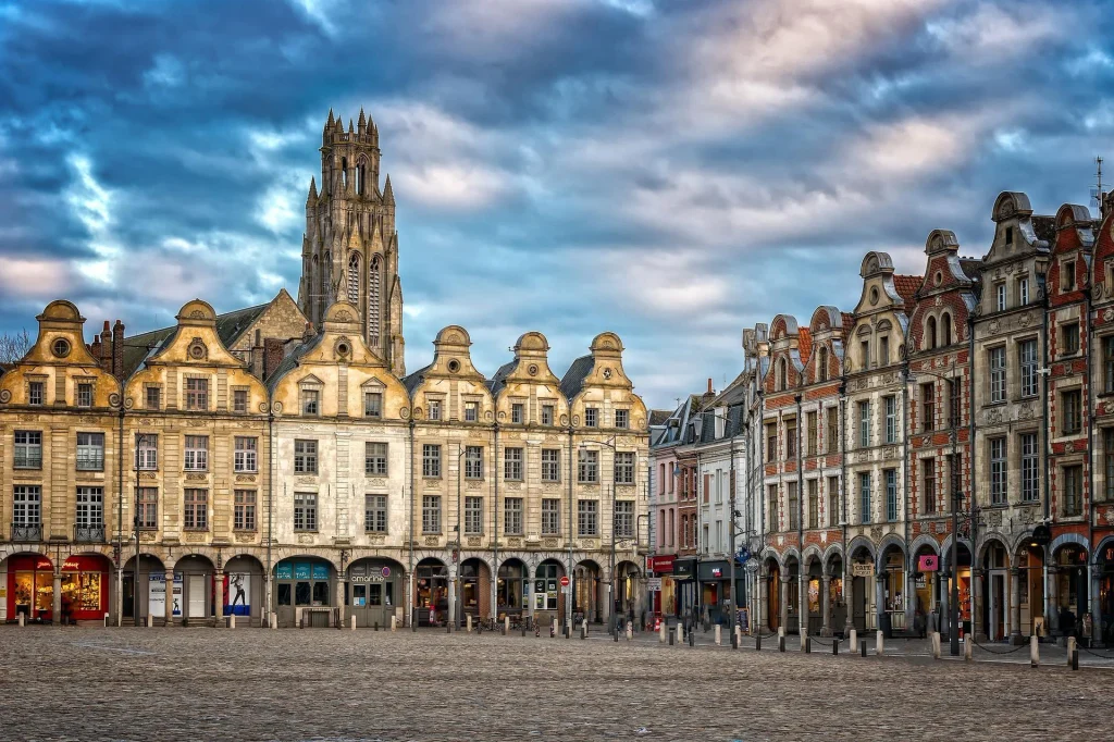 Historic square in Arras, northern France, featuring Flemish-Baroque architecture and a Gothic church tower beneath a dramatic sky.