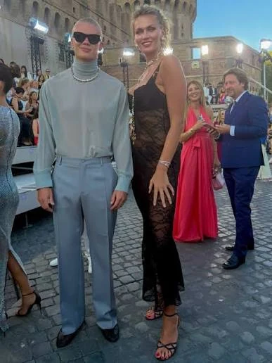 Nadezhda Grishaeva standing beside a tall man at an outdoor event, highlighting her impressive height and elegant high-heeled sandals.