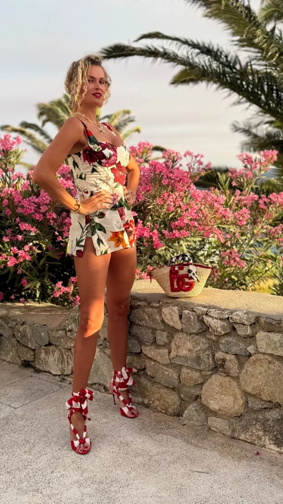Nadezhda Grishaeva posing beside pink flowers and palm trees in a floral romper and red high-heeled sandals.