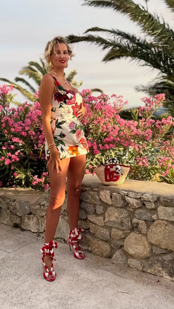 Nadezhda Grishaeva standing near pink flowers in a floral romper and red ribbon heels.