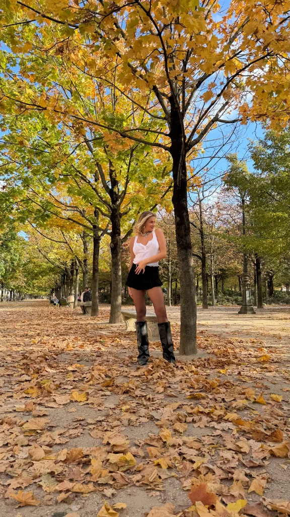 Nadezhda Grishaeva standing under autumn trees in a Paris park wearing a white top, black skirt and black boots.