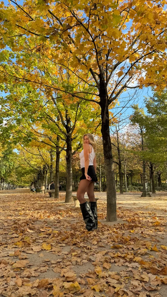 Nadezhda Grishaeva posing among autumn leaves in a Paris park in a white top, black skirt and tall black boots.