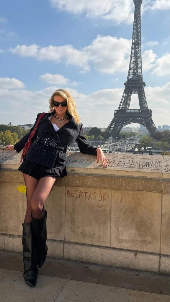 Nadezhda Grishaeva smiling in front of the Eiffel Tower in Paris in a black belted outfit and tall boots.